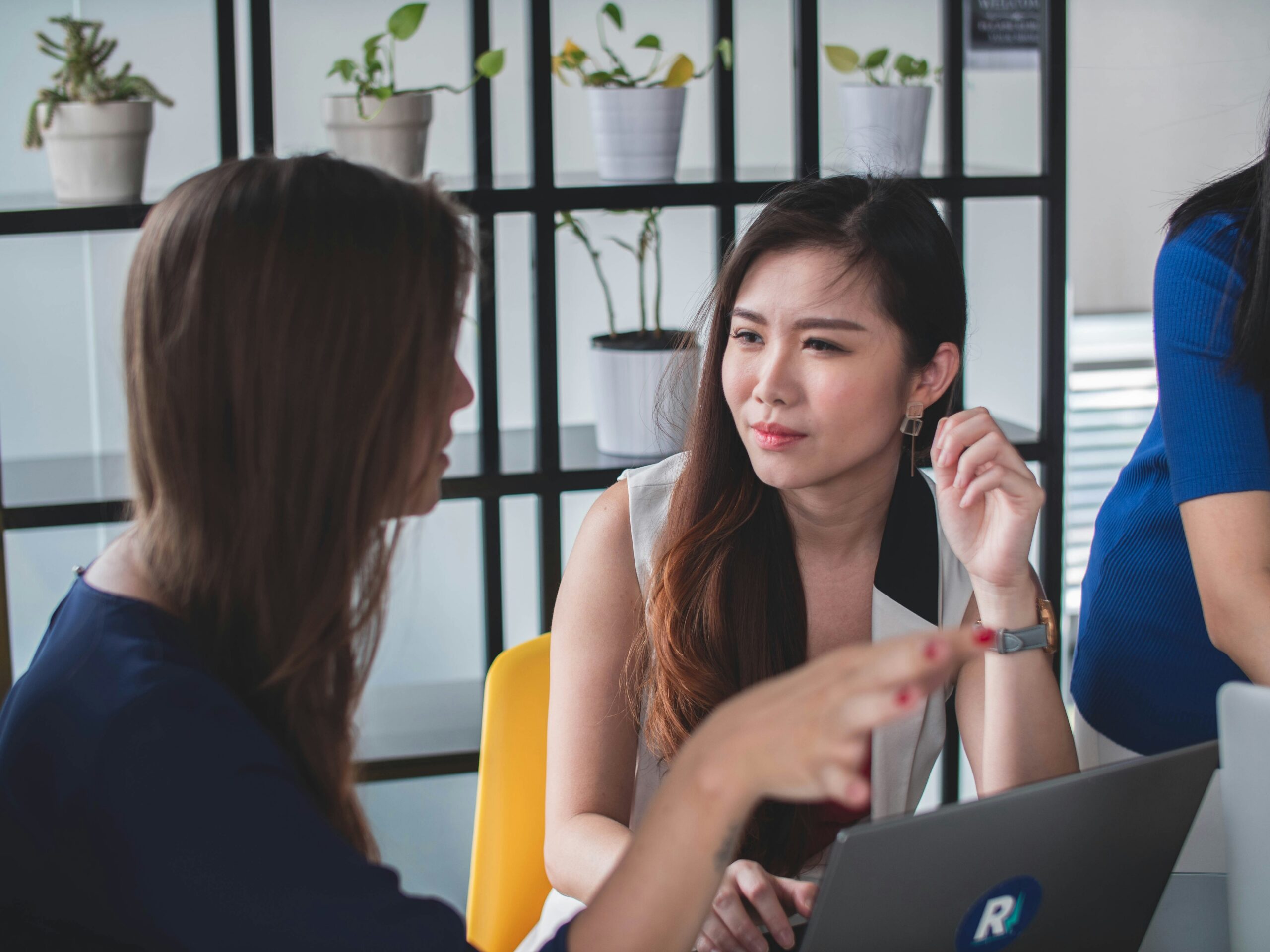 Two women talking at a desk with a laptop, plants on a grid partition behind them.
